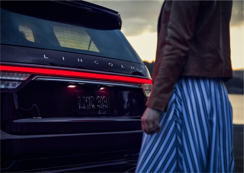 A person is shown near the rear of a 2023 Lincoln Aviator® SUV as the Lincoln Embrace illuminates the rear lights | Star Lincoln in Southfield MI